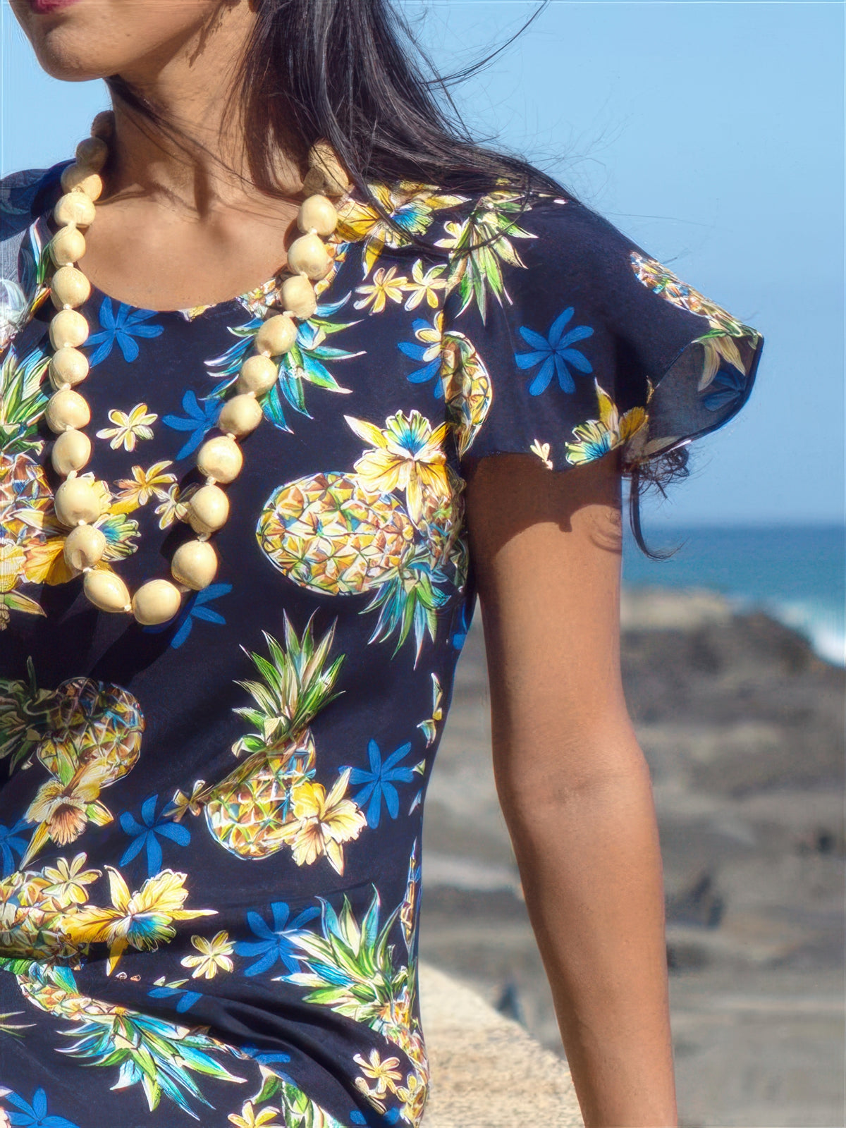 Woman wearing a colorful pineapple-patterned dress with a necklace, standing against a beach backdrop.