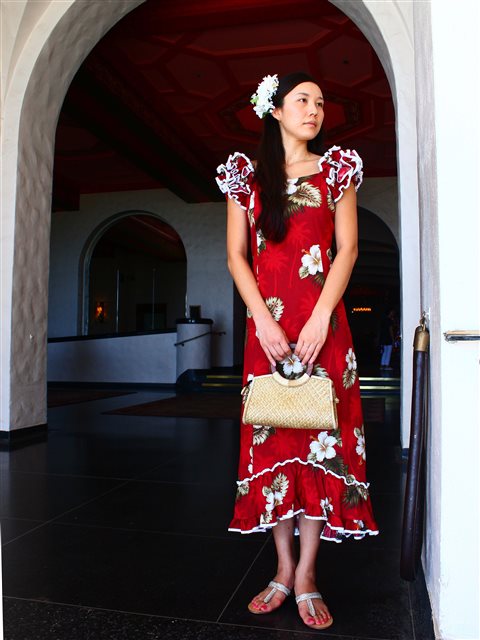 A woman standing in a indoor setting wearing a red and navy floral Hawaiian muumuu dress with ruffle sleeves and a frill skirt hem. She has a flower in her hair and is holding a beige purse.