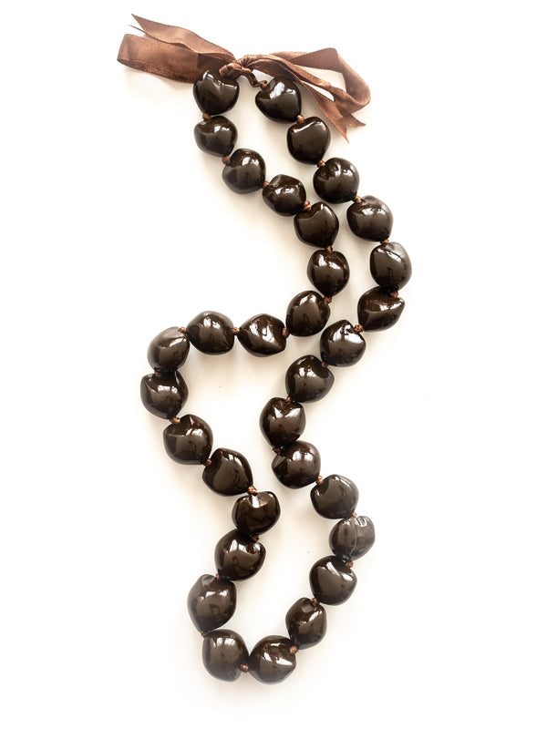 A brown kukui nut lei with a ribbon tied at the end, laid out on a white background.